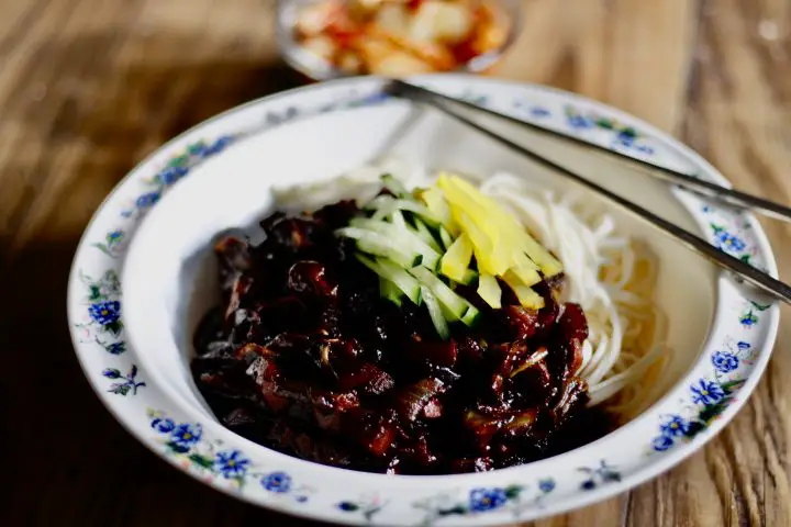 A white bowl with blue flowers on the edge holding jajangmyeon topped with cucumber and pickled daikon matchsticks with metal chopsticks and kimchi in the background