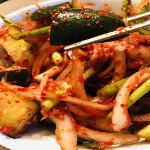 Cucumber kimchi in a white bowl with silver chopsticks holding up a cucumber