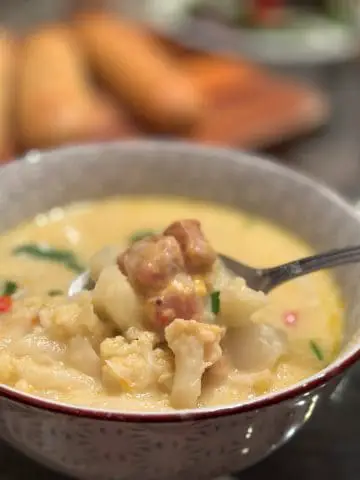 Creamy Cauliflower, Potato, and Cheesy Ham Chowder in a bowl with a spoon holding some of the chowder and breadsticks in the background.
