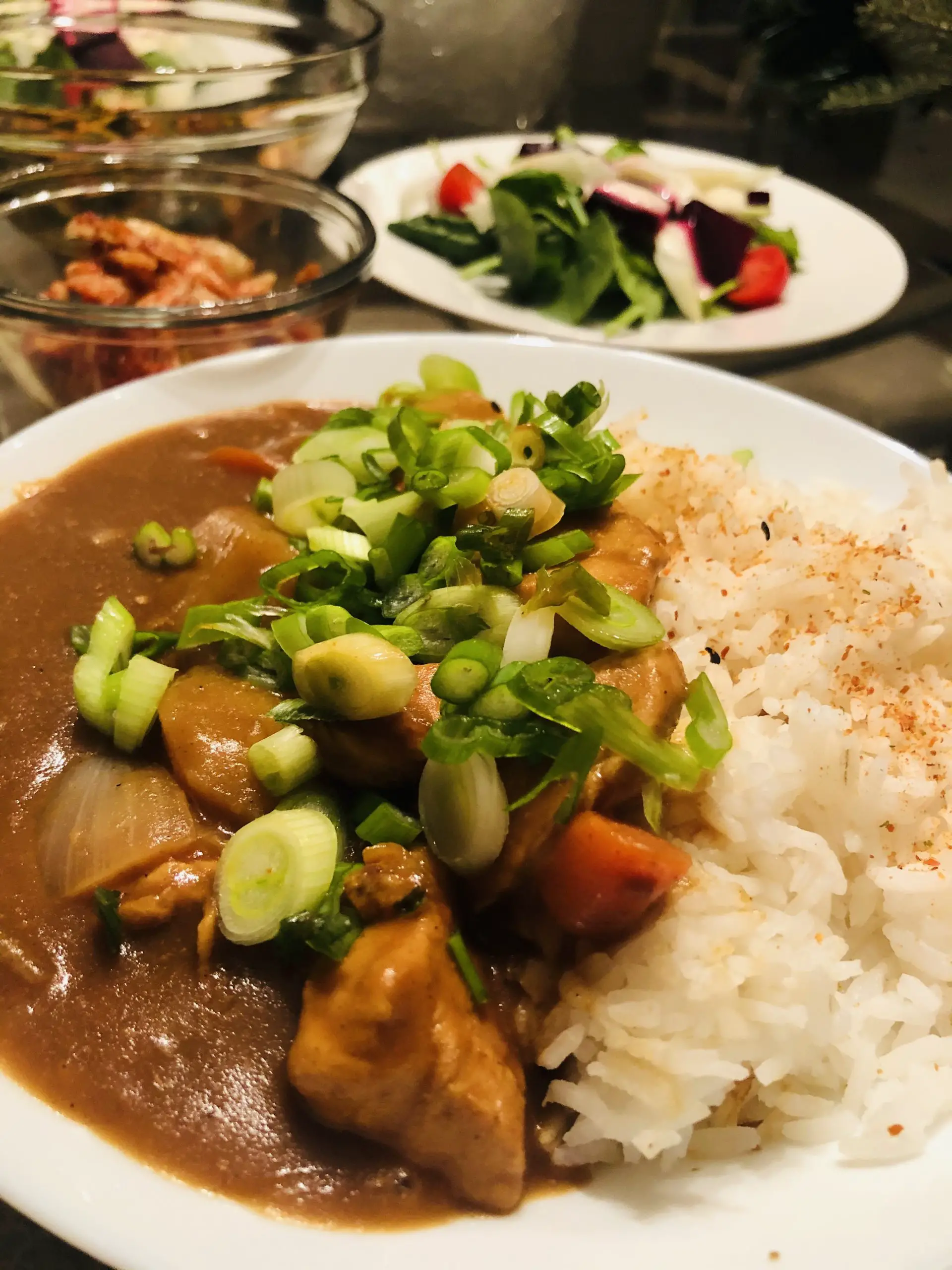 Vermont Curry With Chicken served with rice and garnished with green onions, with kimchi and a salad in the background