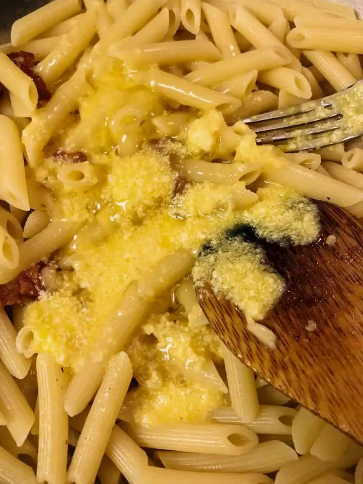 Penne pasta with whisked egg and Parmesan cheese.  There is a wooden spoon and a fork resting in the pasta. 