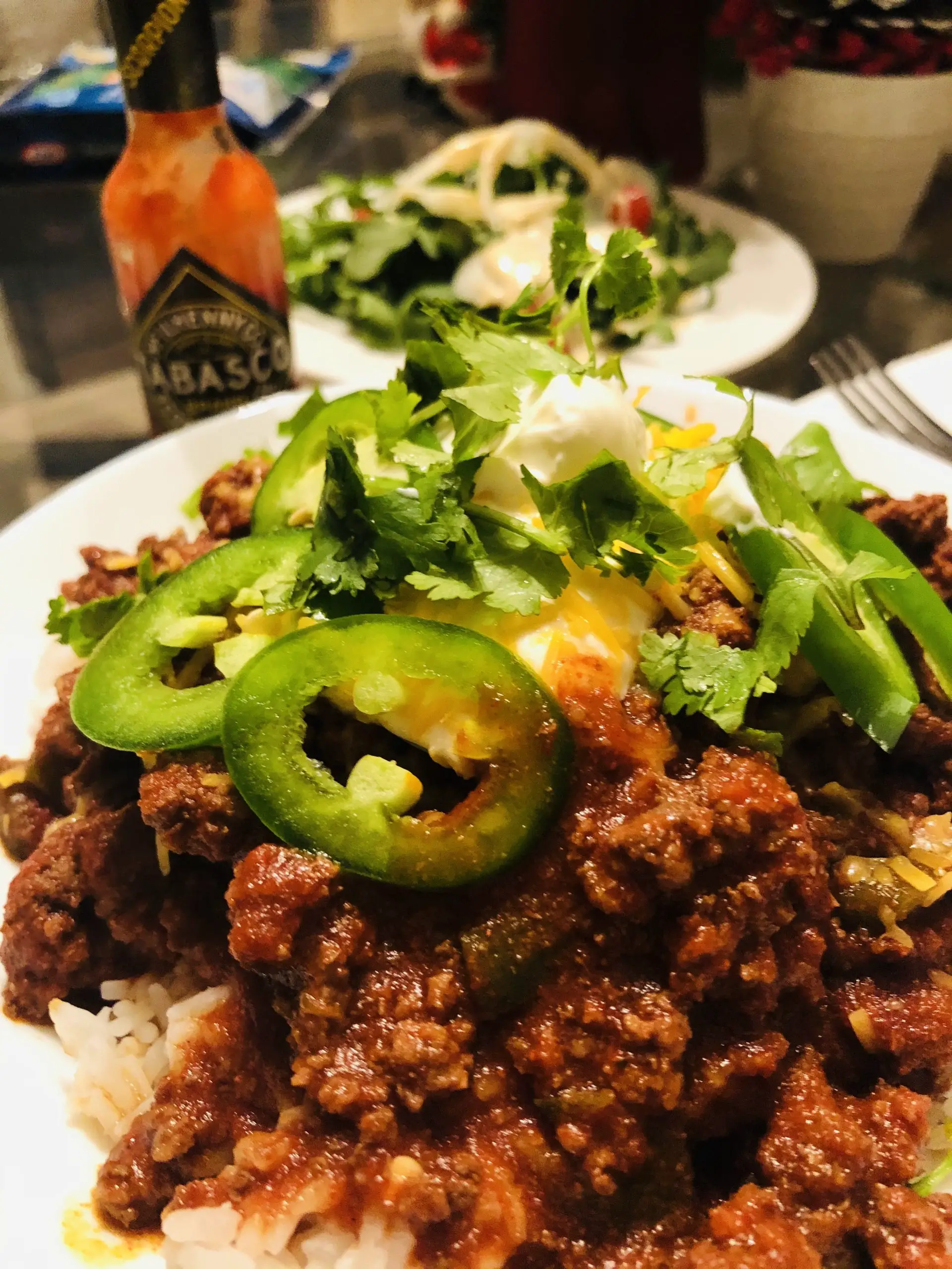 Texas Chili with sliced jalapenos, sour cream, cilantro, and cheese with a Tabasco bottle and salad in the background served with rice