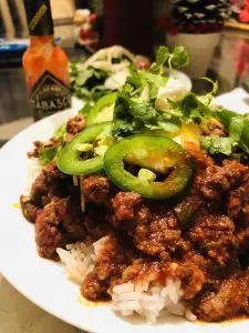 Texas Chili with sliced jalapenos, sour cream, cilantro, and cheese with a Tabasco bottle and salad in the background served with rice