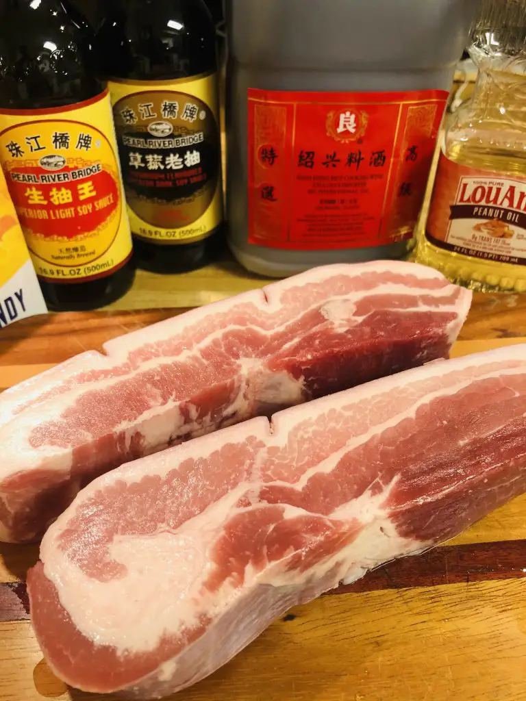 2 slabs of pork belly on a wooden cutting board, and behind that are bottles of peanut oil, Shaoxing rice wine, dark soy sauce, light soy sauce, and rock candy