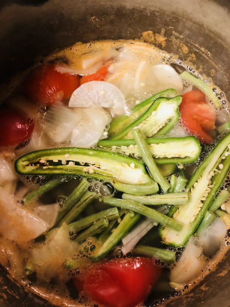veggies including sliced serrano peppers, tomatoes, onion, and radish with Mama Sita's sinigang seasoning in a saucepan with water