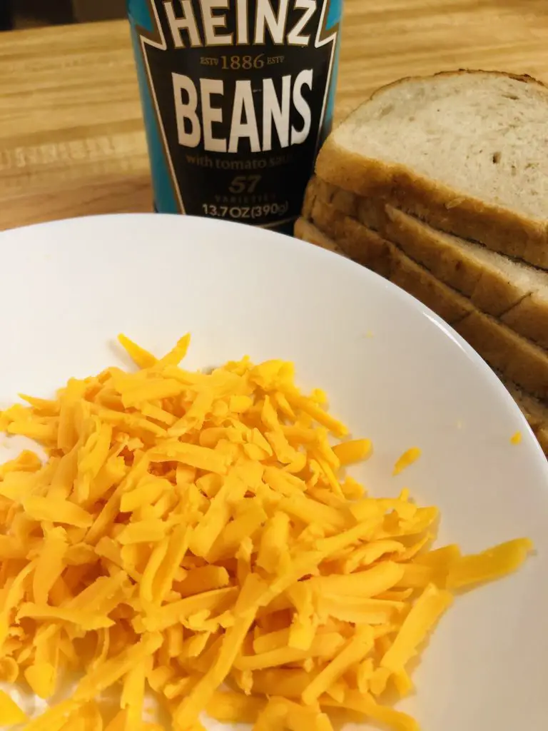 grated cheddar cheese in a white bowl, with slices of sourdough and Heinz beans in the background