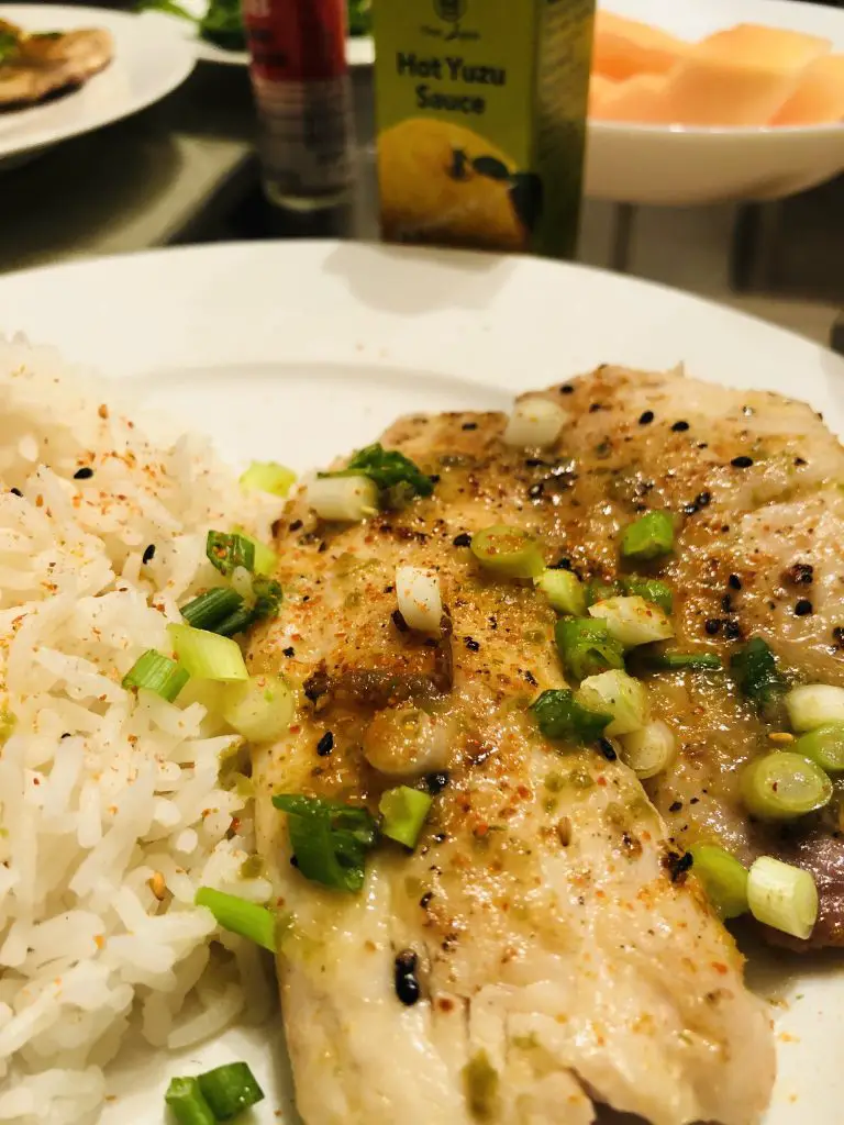 Tilapia With Hot Yuzu Sauce and rice garnished with green onions on a white plate with Hot Yuzu Sauce and cut up cantaloupe in a white bowl in the background