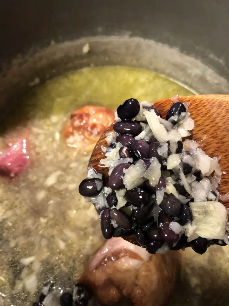 black beans, minced garlic and onion, broth and ham hocks in a saucepan, with minced onion and black beans on a wooden spoon in the foreground