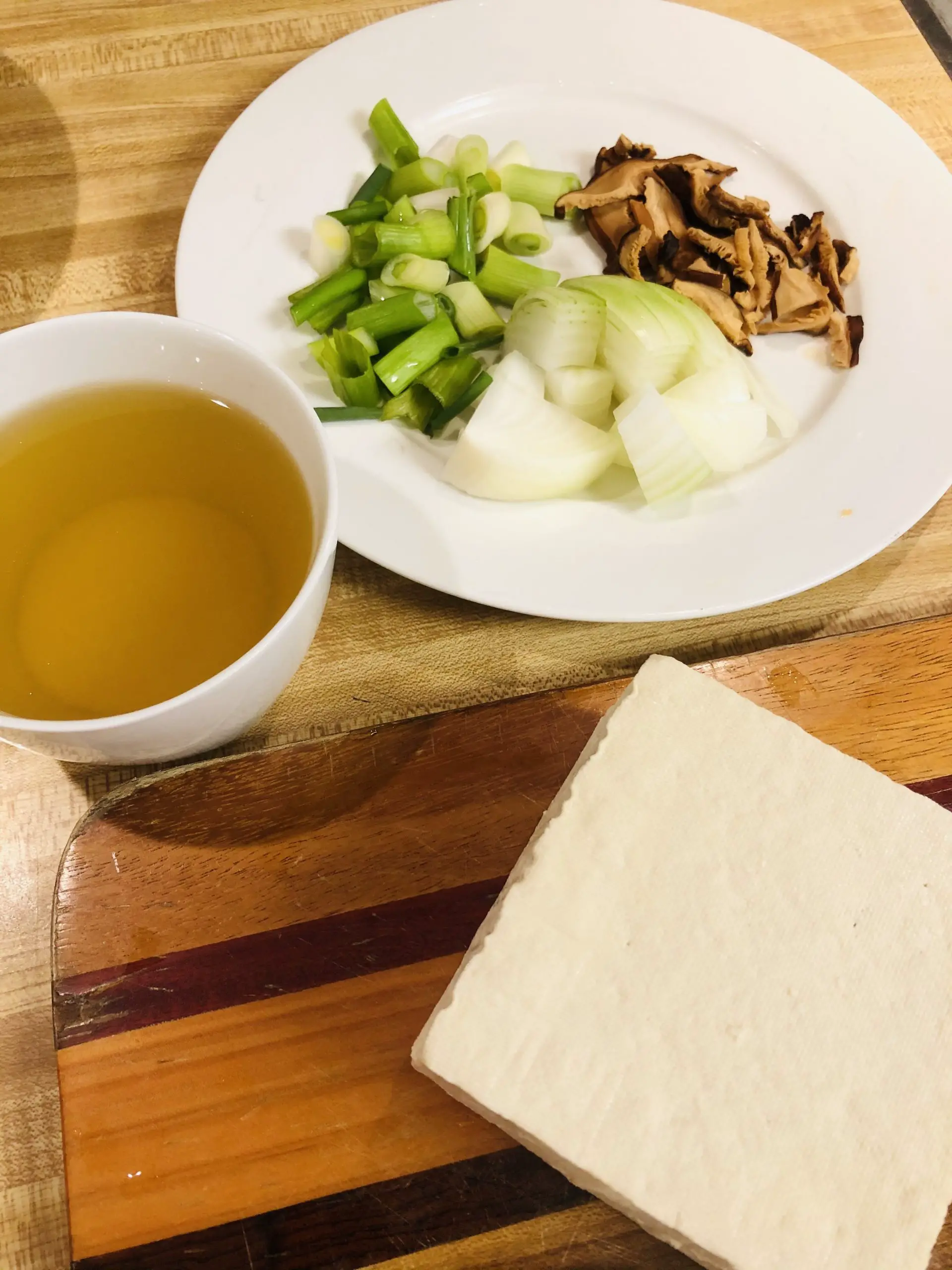 anchovy broth in a white bowl, block of soft tofu, green onions, onions, and shiitake mushrooms on a white plate