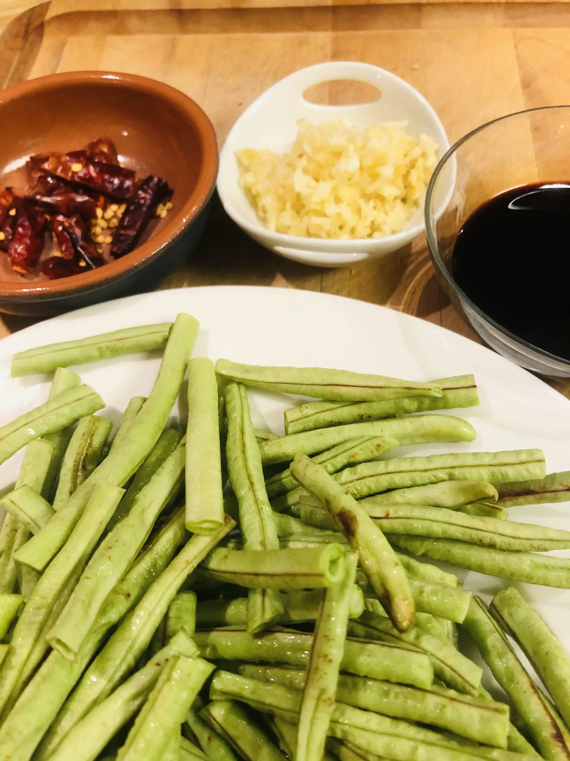 Chinese long beans cut into 3 inch pieces on a white plate, ground up Sichuan peppercorn in a small bowl, minced ginger and garlic in a small bowl, and a glass bowl with soy sauce and shaoxing rice wine
