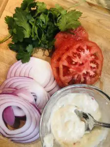 Mayo with Thai chilis, fish sauce and garlic in a small bowl, cilantro leaves, sliced tomatoes, and sliced onions, on a wooden bowl