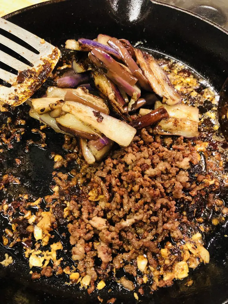 stir fried eggplant and crispy minced pork added to seasonings in a cast iron pan with spatula on the side