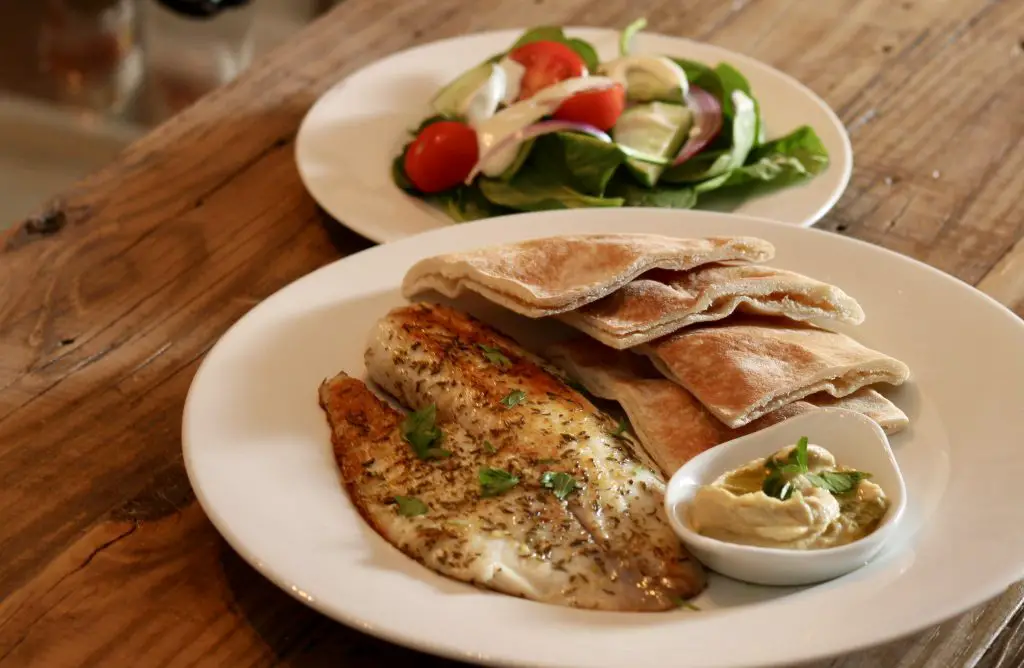 Tilapia With Za'atar garnished with parsley, pieces of pita bread, and hummus on a white plate with salad on a white plate behind the plate with the tilapia