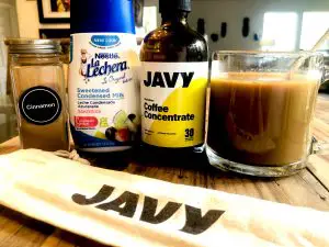 Javy coffee concentrate bottle alongside a glass filled with cold coffee, a glass bottle with cinnamon, a bottle of sweetened condensed milk, and a cloth labeled with Jafy in front of these items