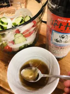 Cucumber salad veggies in a glass bowl with a bottle of fish sauce and a bowl of Thai Cucumber salad sauce with a spoon