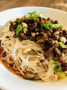 Dan dan noodles topped with pork mixture, scallions and peanuts in a white bowl