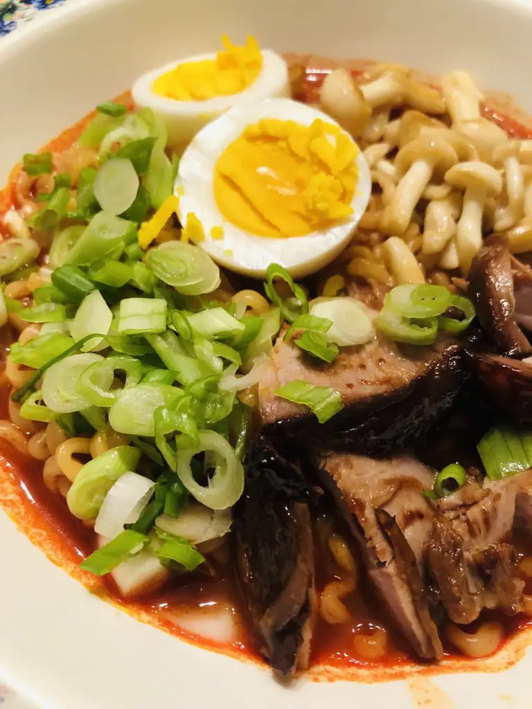 Asian noodles being held up by chopsticks;  soup in a bowl with hoisin pork, mushrooms, eggs, and green onions