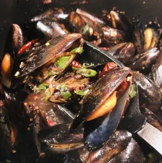 Mussels With Black Bean Sauce garnished with green onions and dried Asian chilies, with a spoon holding some of the mussels