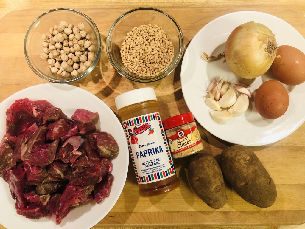 beef stew cuts in a white bowl, paprika, ground ginger, two potatoes, glass bowls of barley and chickpeas, a white bowl containing an onion, two eggs, and cloves of garlic