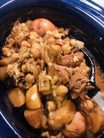 Cholent in a slow cooker with a black spoon scooping up some of the cholent