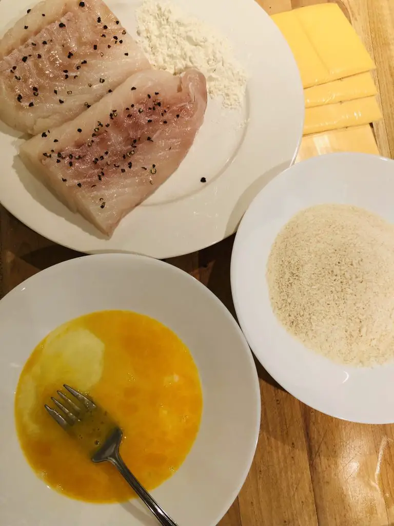 Cod loins and flour on a white plate, whisked egg and fork in a white bowl, panko in a white bowl, and slices of American cheese