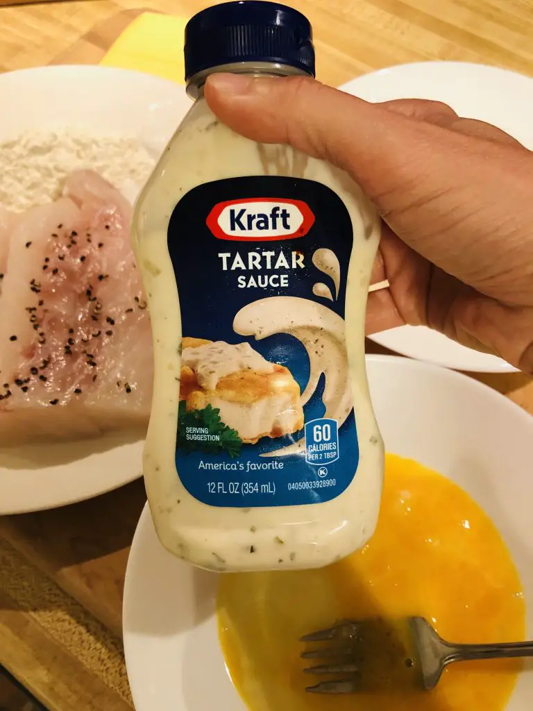 A hand holding a bottle of Kraft Tartar sauce with cod and whisked egg in the background