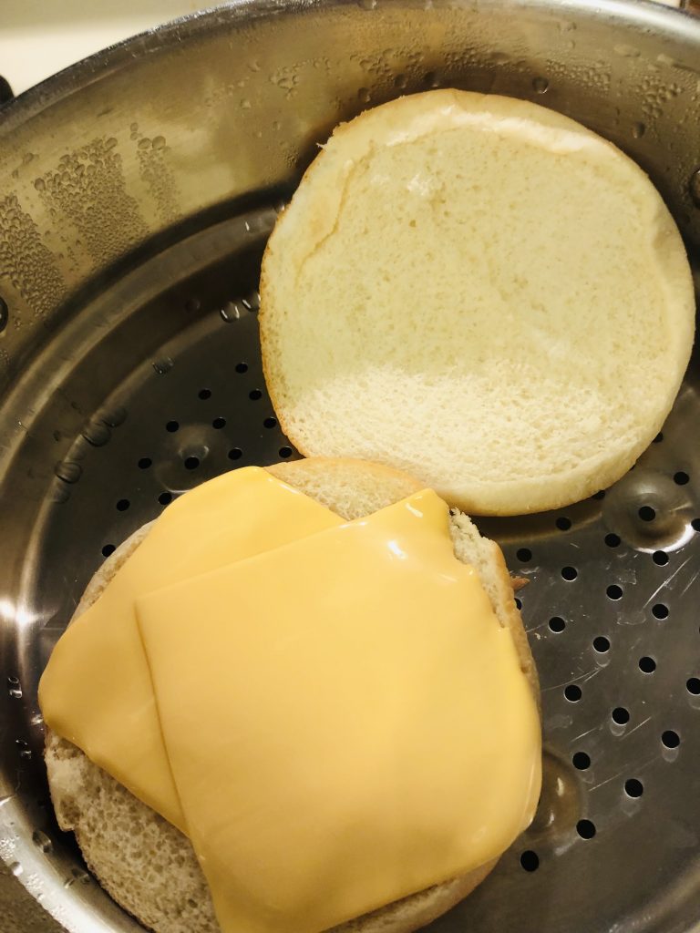 Burger buns in a steam bath with American cheese slices on the bottom half