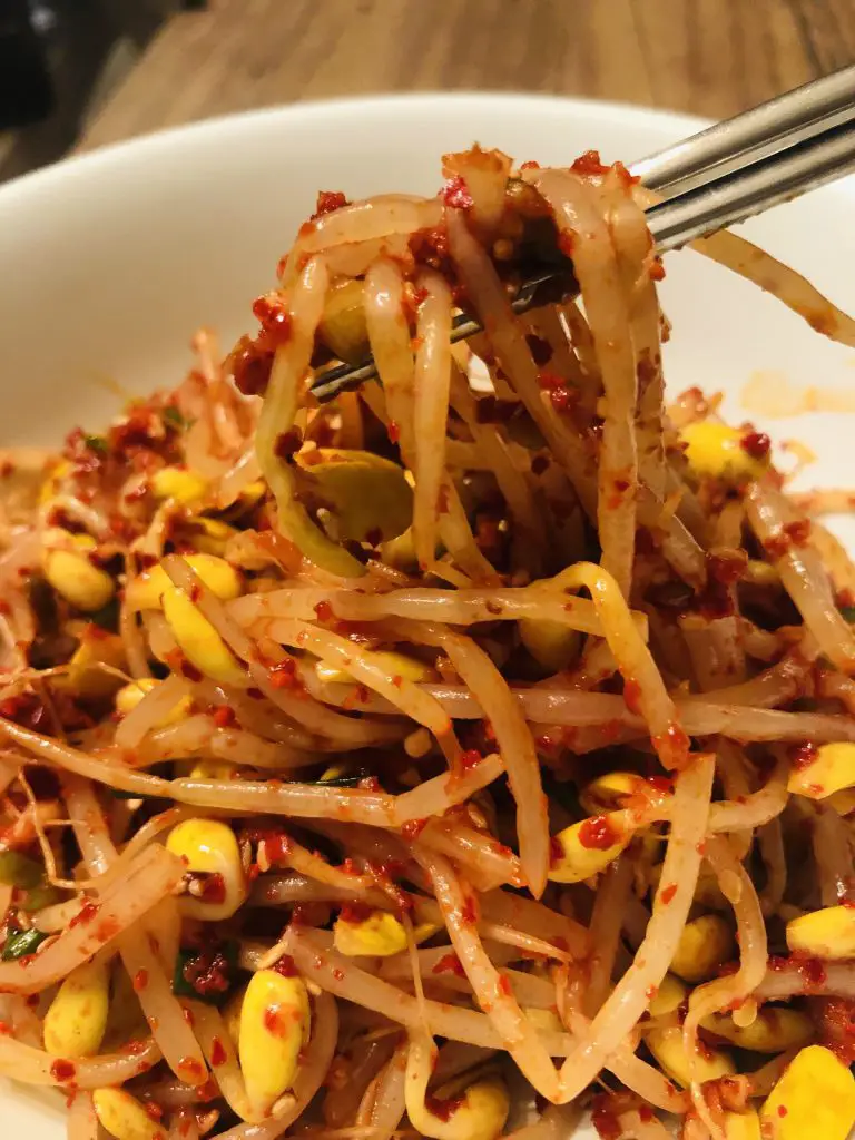 Korean Spicy Bean Sprouts in a white bowl with some silver chopsticks holding some of the bean sprouts