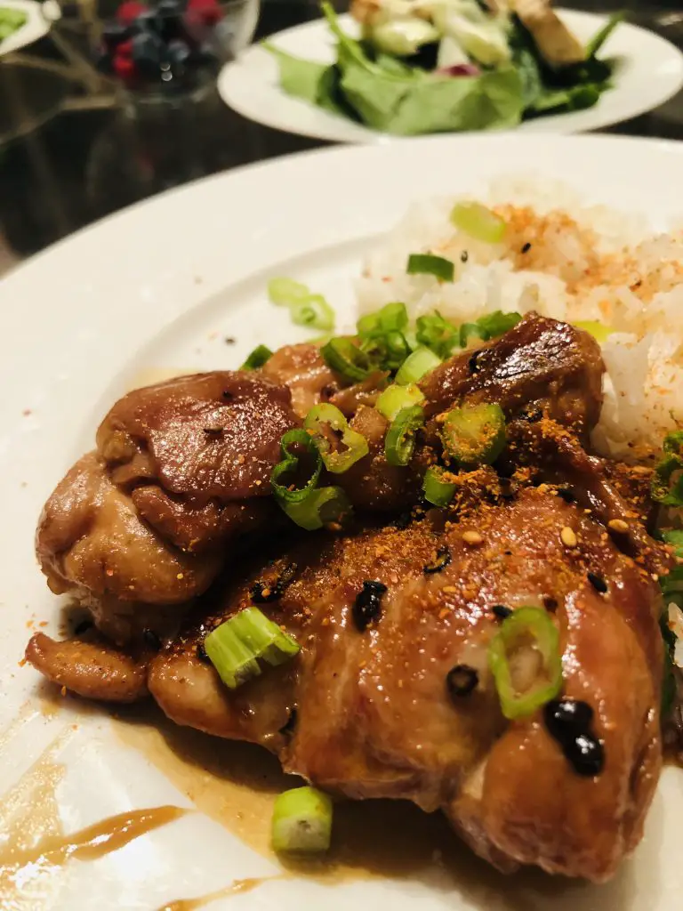 Teriyaki Chicken garnished with green onions and Nanami Togarashi served with rice on a white plate, with a salad and berries in the background