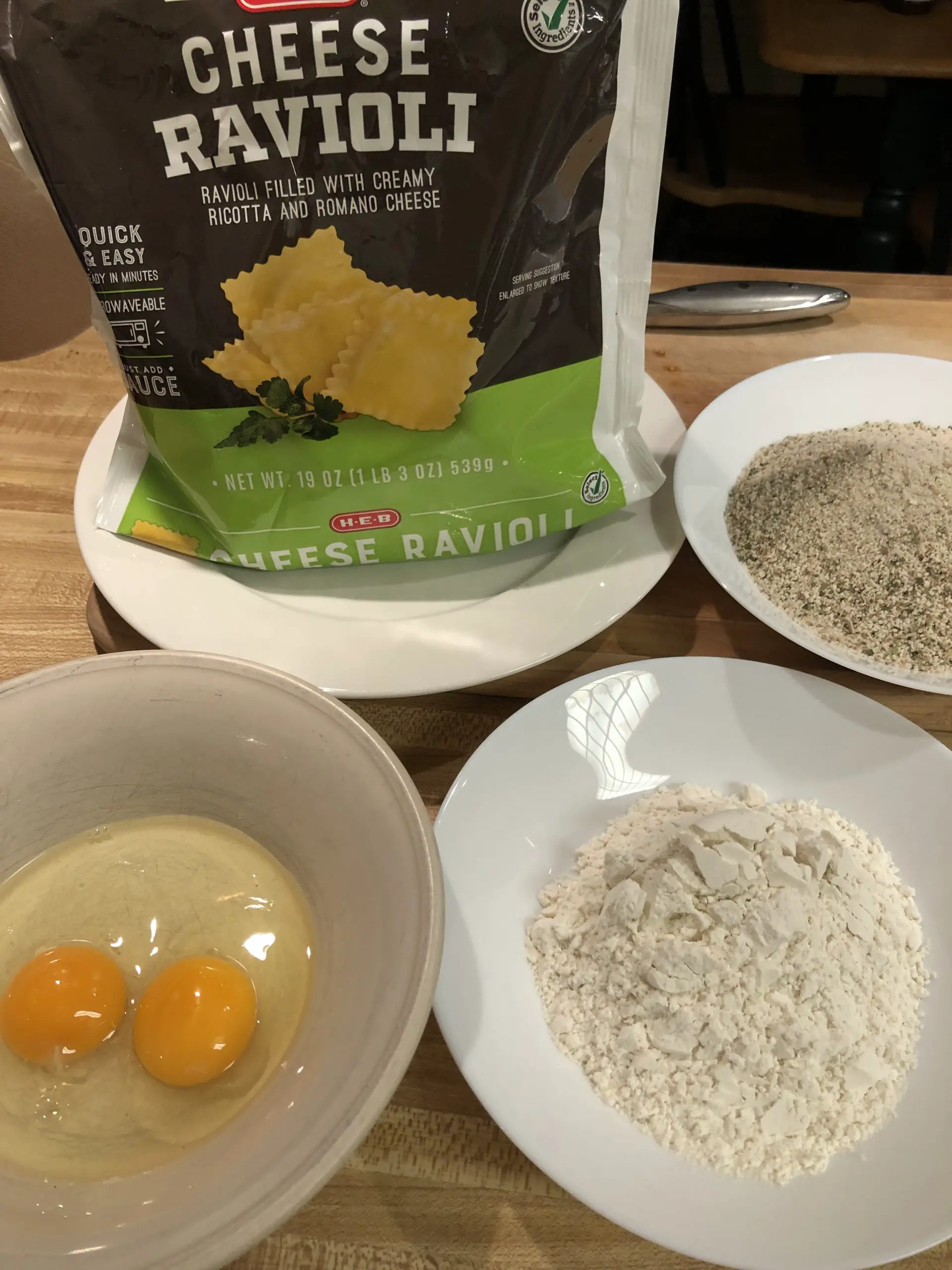 Bag of cheese ravioli, eggs in a bowl, flour in a white bowl, Italian breadcrumbs in a white bowl