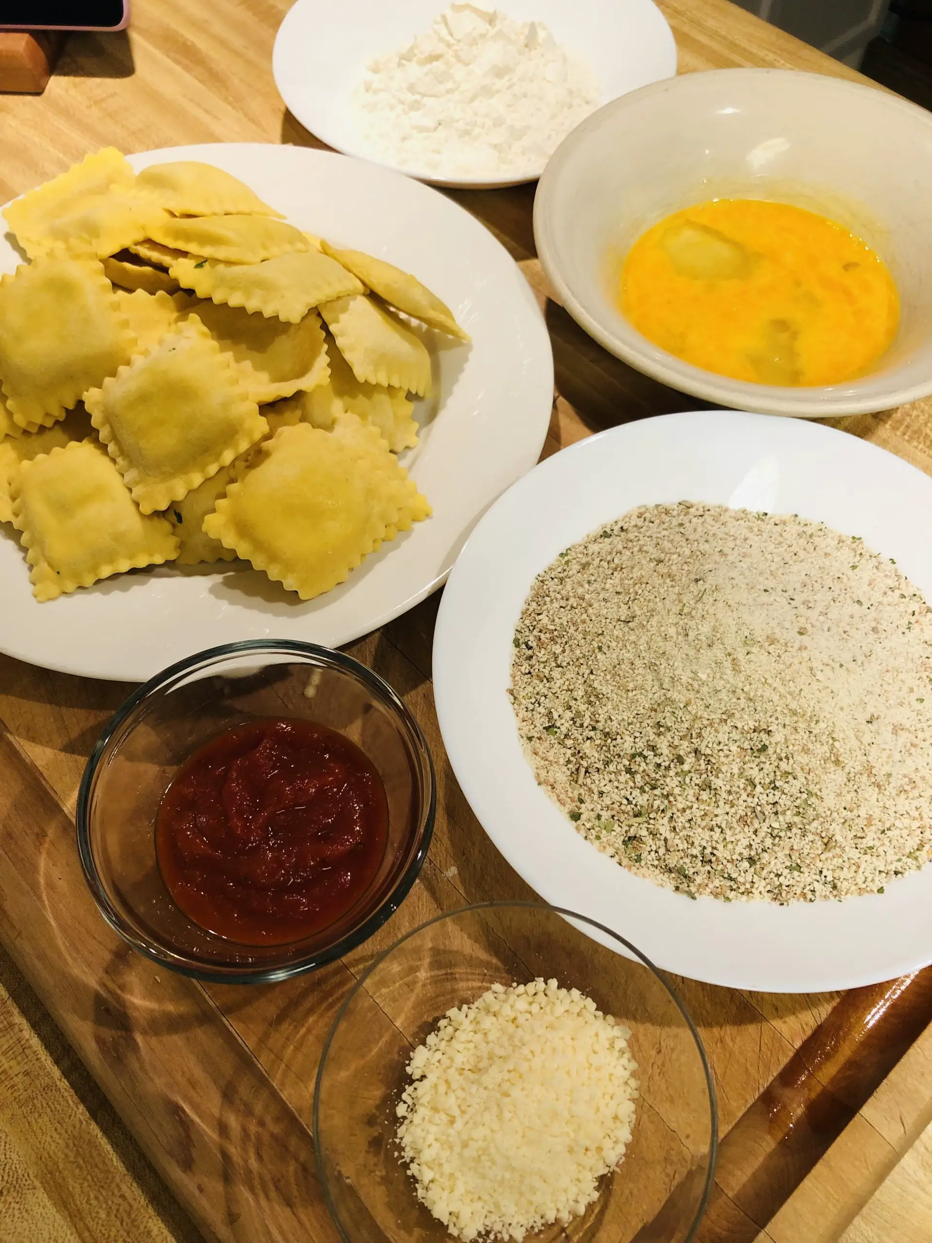 Ravioli on a white plate, whisked eggs in a bowl, Italian breadcrumbs in a white bowl, flour in a white bowl, marinara sauce and parmesan cheese in small glass bowls