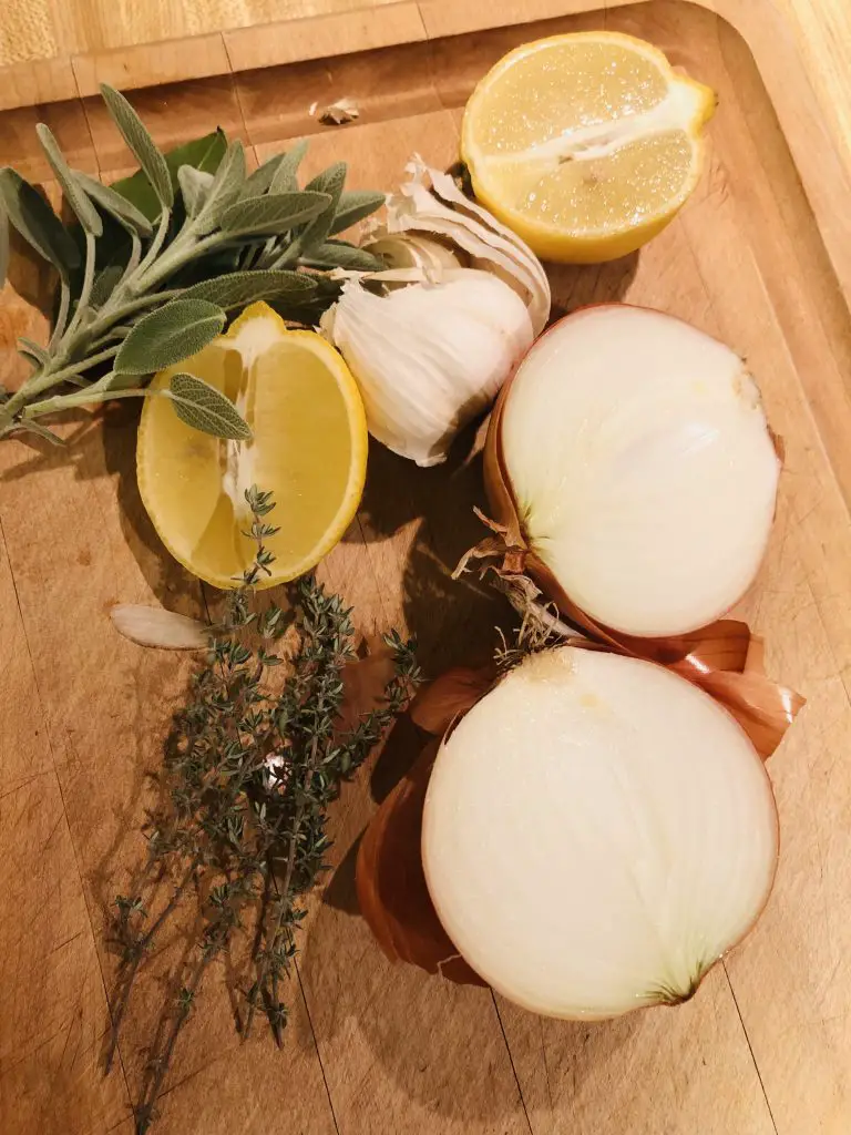 halved lemon and onion, thyme and sage, and some cloves of garlic on a wooden board 
