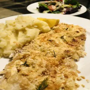 Oven baked fish with mashed potatoes and a lemon wedge on a white plate with a salad in the background