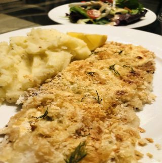 Oven baked fish with mashed potatoes and a lemon wedge on a white plate with a salad in the background