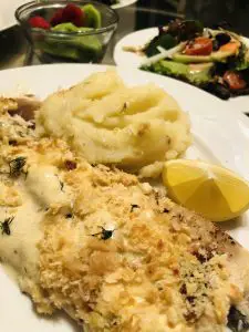 Oven baked fish with mashed potatoes and a lemon wedge on a white plate with a salad and fruit in the background