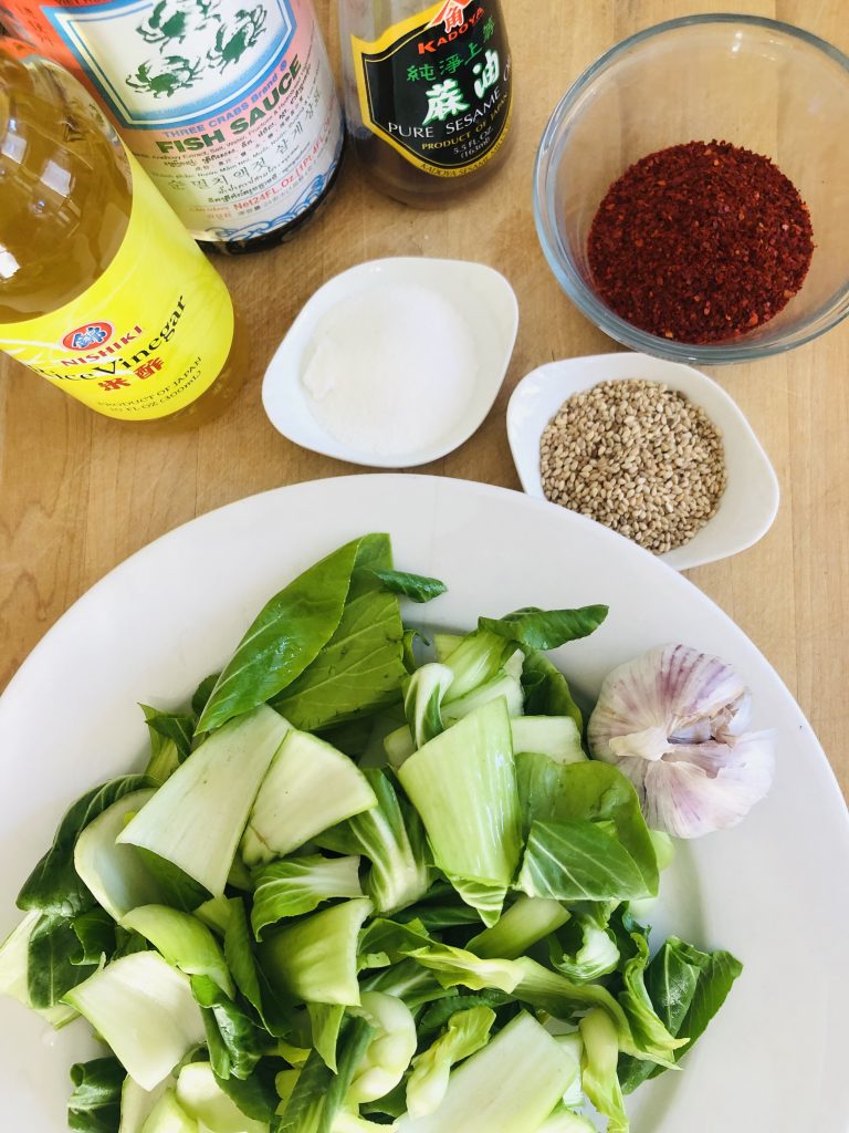 cut up pieces of bok choy on a white plate, sugar, sesame seeds, and Korean red pepper powder in small bowls, bottles of rice vinegar, fish sauce, and sesame oil