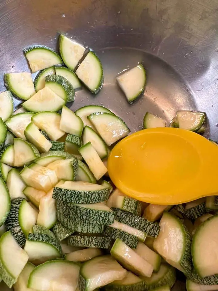 A silver bowl containing slices of zucchini and water drawn out from the zucchini. There is a yellow spoon in the bowl.