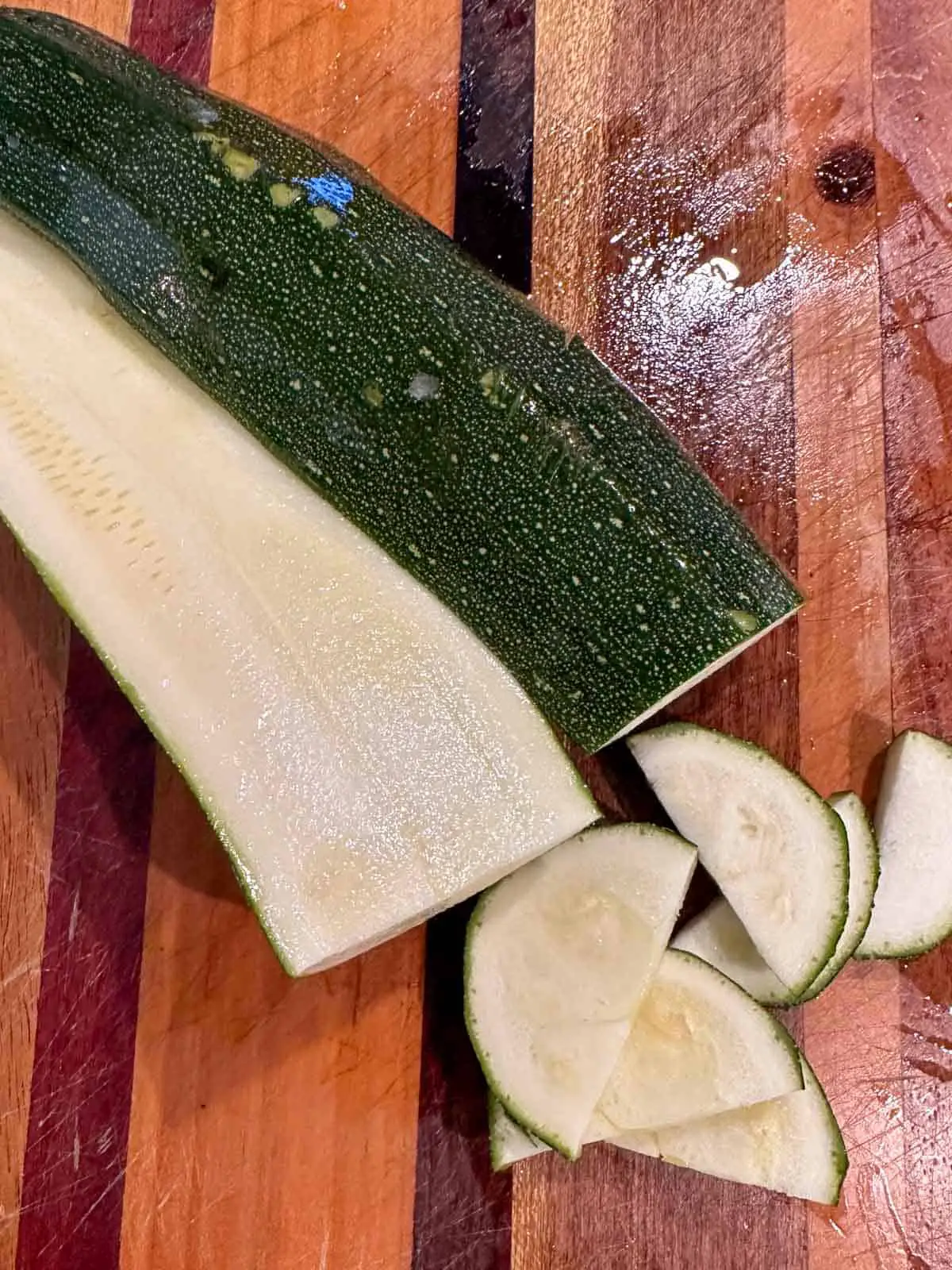 A zucchini on a wooden cutting board which has been sliced down the middle, then a few slices were cut across into half moon shapes.