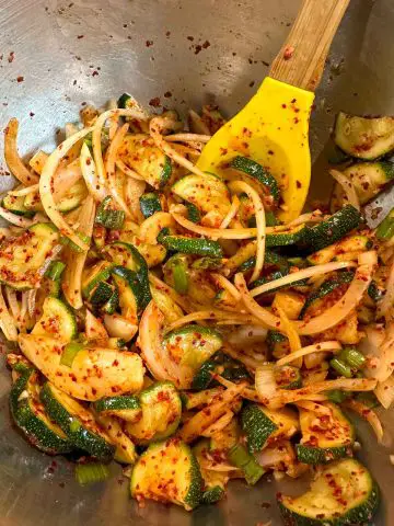 Zucchini kimchi which is slices of zucchini, sliced onion and green onions in a spicy red kimchi sauce. There is a yellow spoon with a wooden handle resting in the bowl.