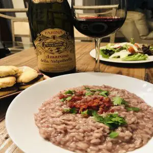 Amarone Red Wine Risotto in a white dish with a bottle of Amarone and glass of Amarone next to it with salad in the background and chocolate cookies