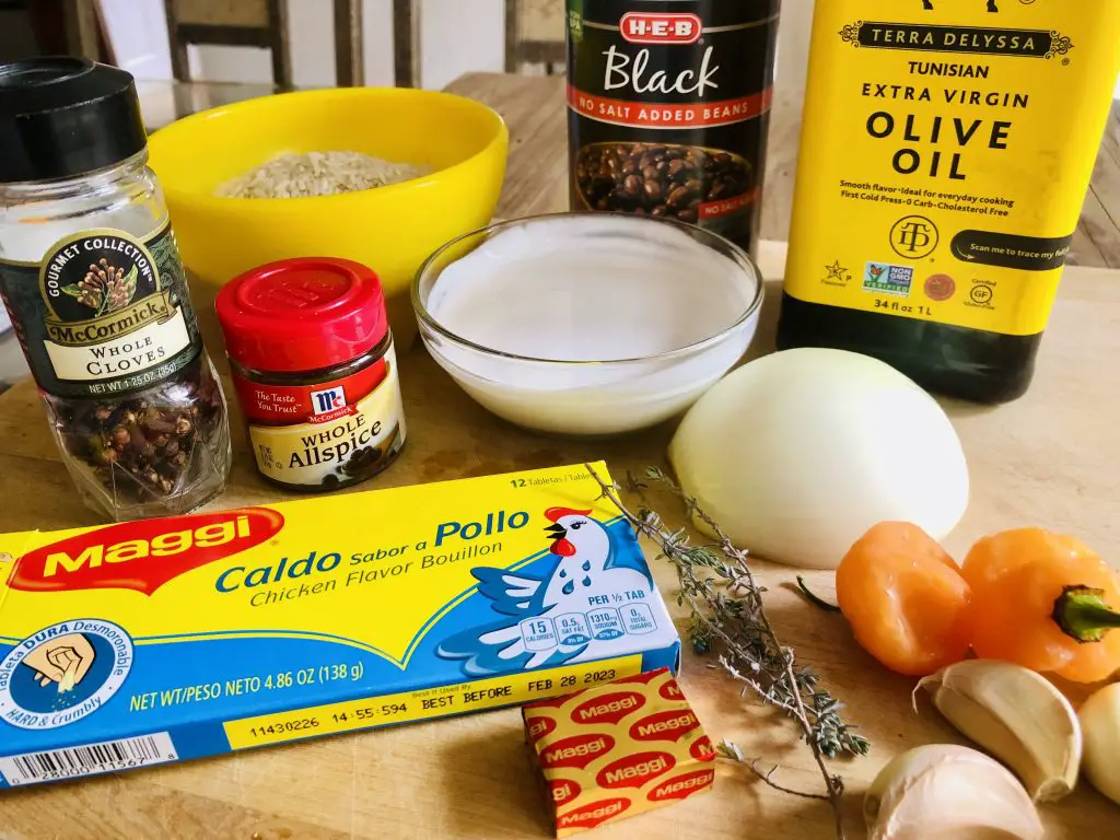 Ingredients for Haitian Rice and Beans including rice, whole cloves, whole allspice, a box of Maggi chicken bouillon and a bouillon cube, sprigs of thyme, garlic, Scotch bonnets, half an onion, coconut milk, can of black beans and bottle of olive oil