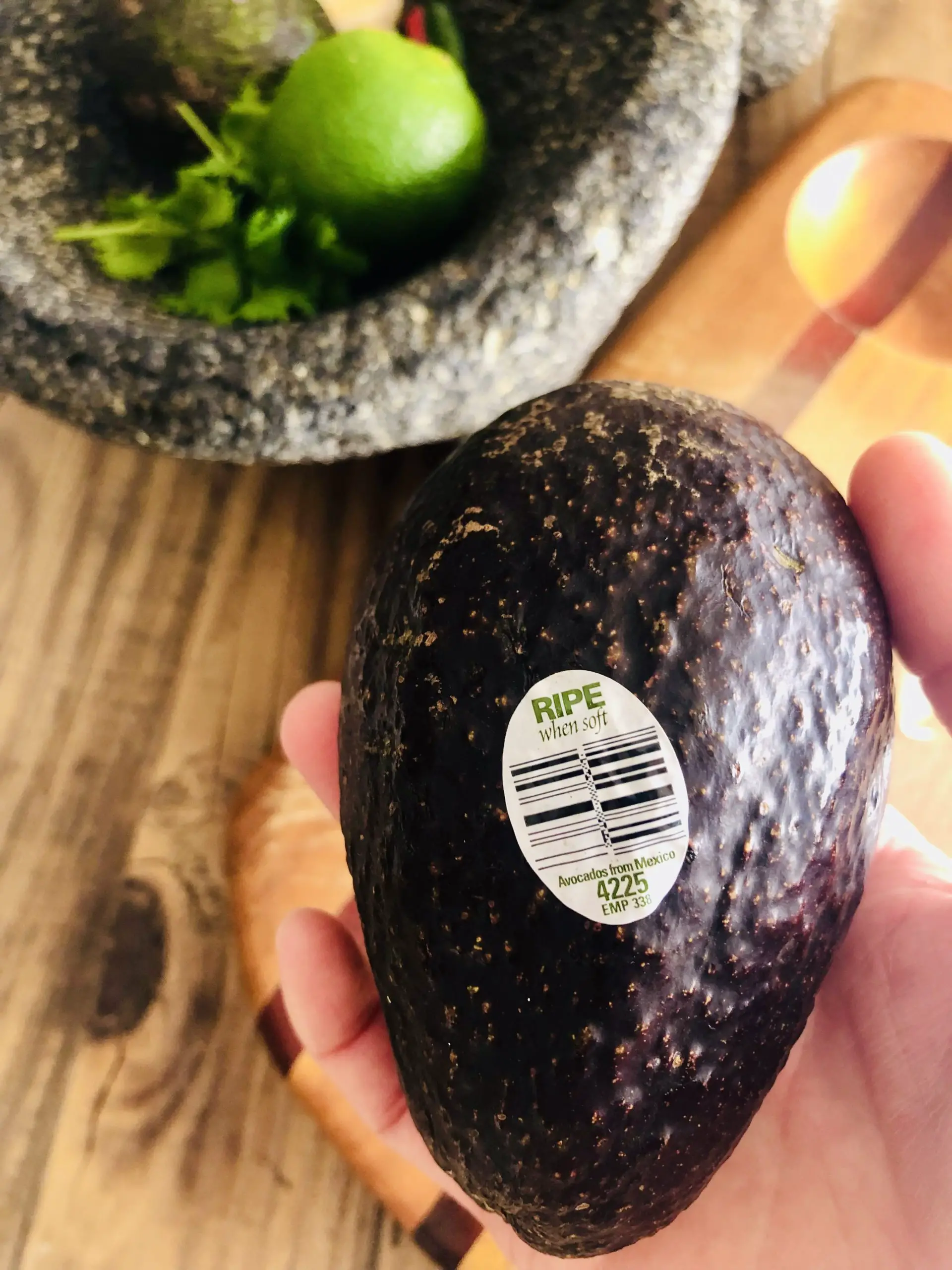 Hand holding an avocado with a sticker that says Ripe when soft, and a molcajete filled with lime, cilantro, and avocado