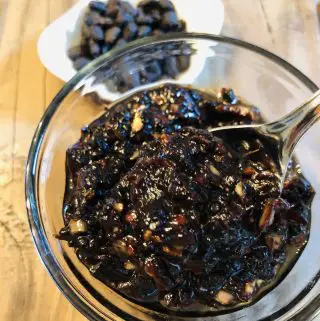 Black bean sauce in a glass bowl with a spoon holding some of the black bean sauce; white dish in the background with fermented black beans