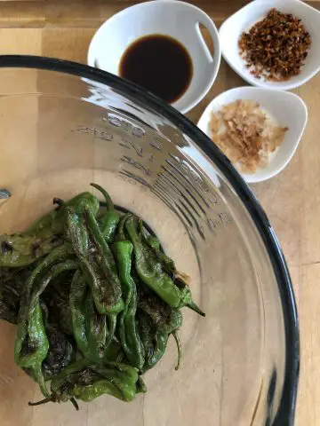 Blistered Shishito peppers in a glass bowl with small white dishes containing soy sauce, furikake, and bonito flakes.