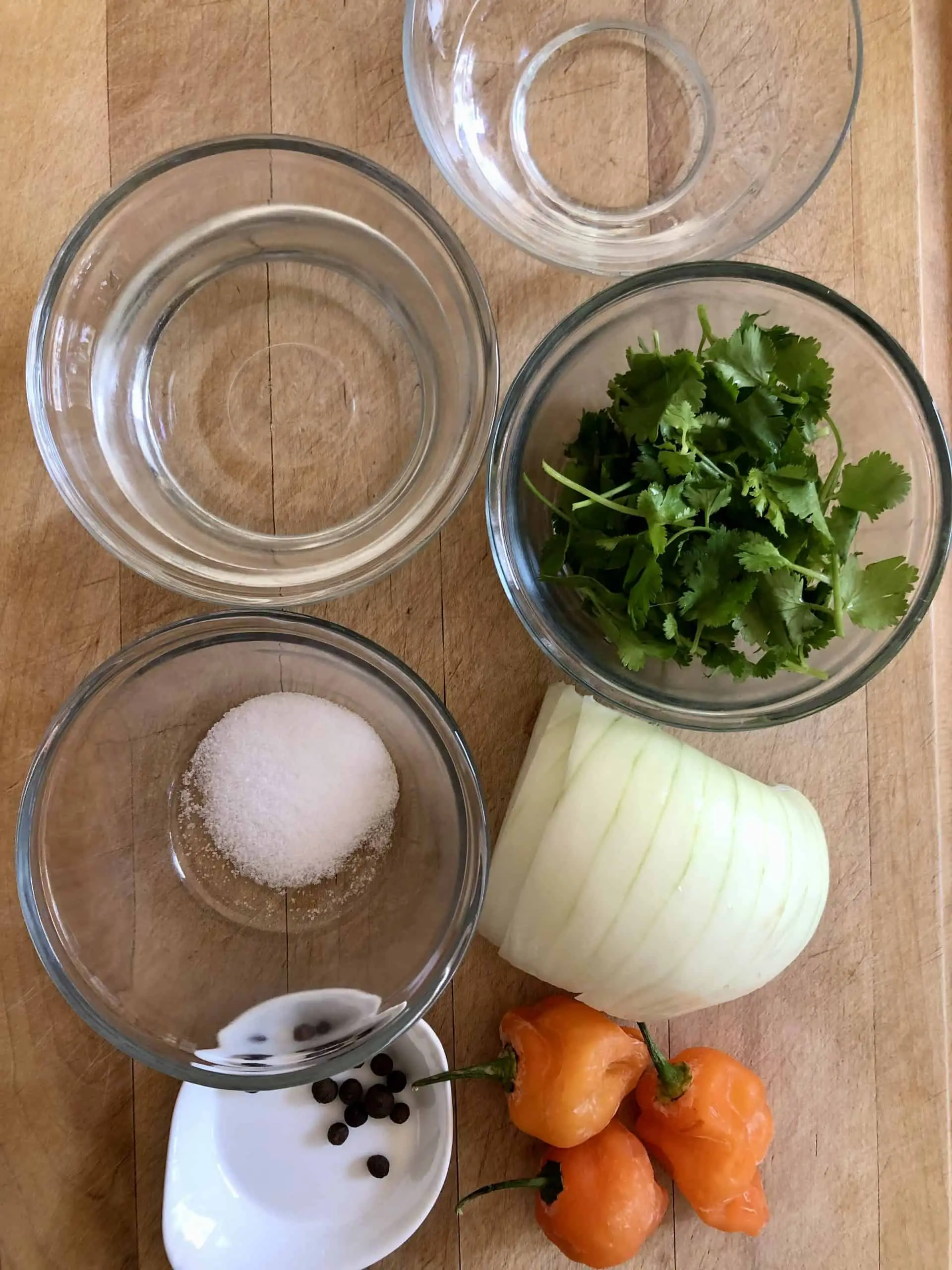 Glass bowls filled with water, vinegar, salt, cilantro, half of an onion, 3 Scotch bonnets, and a small dish with allspice seeds