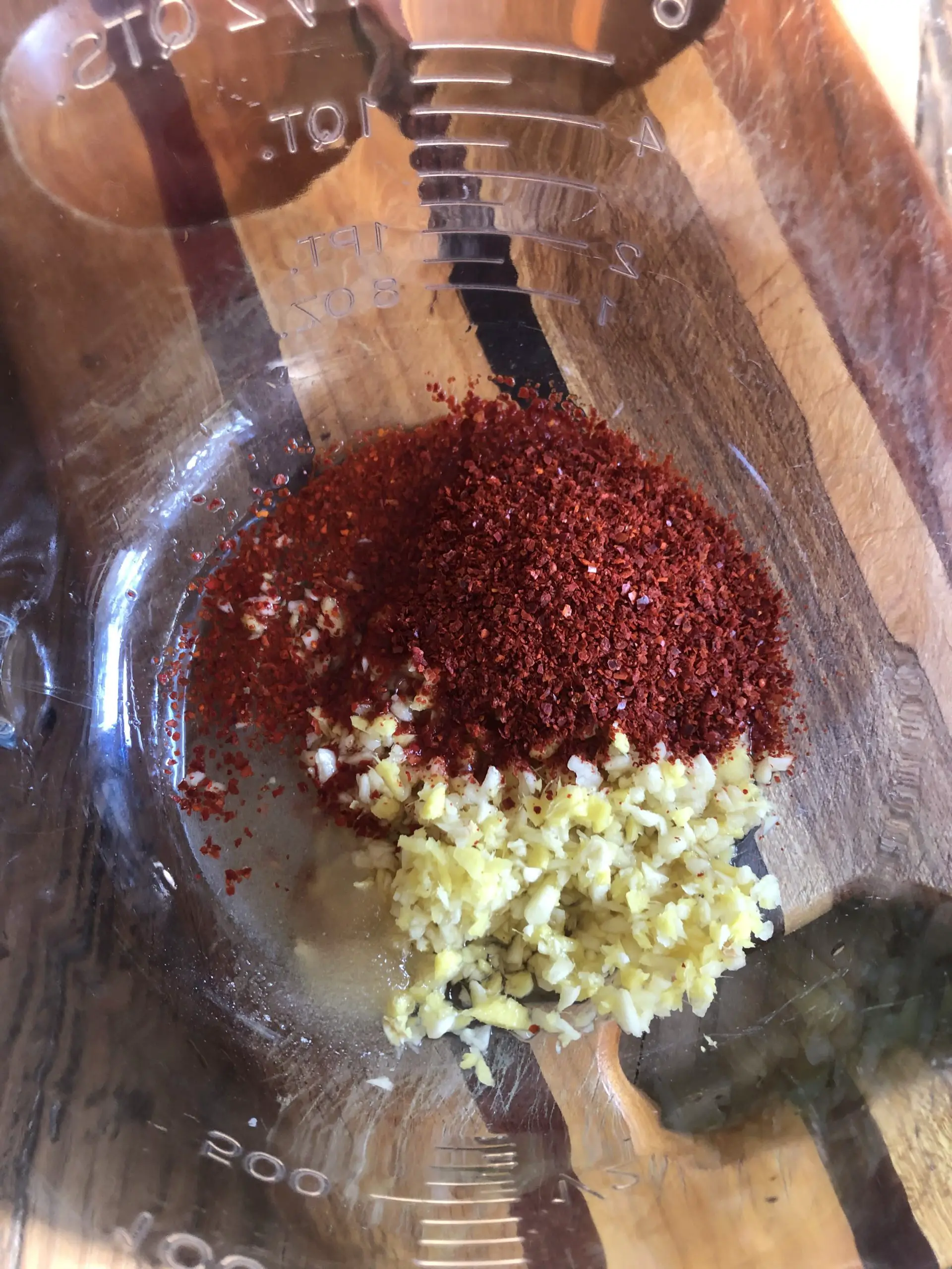 Gochugaru, sauces, and minced ginger and garlic in a glass bowl with a brown cutting board underneath.