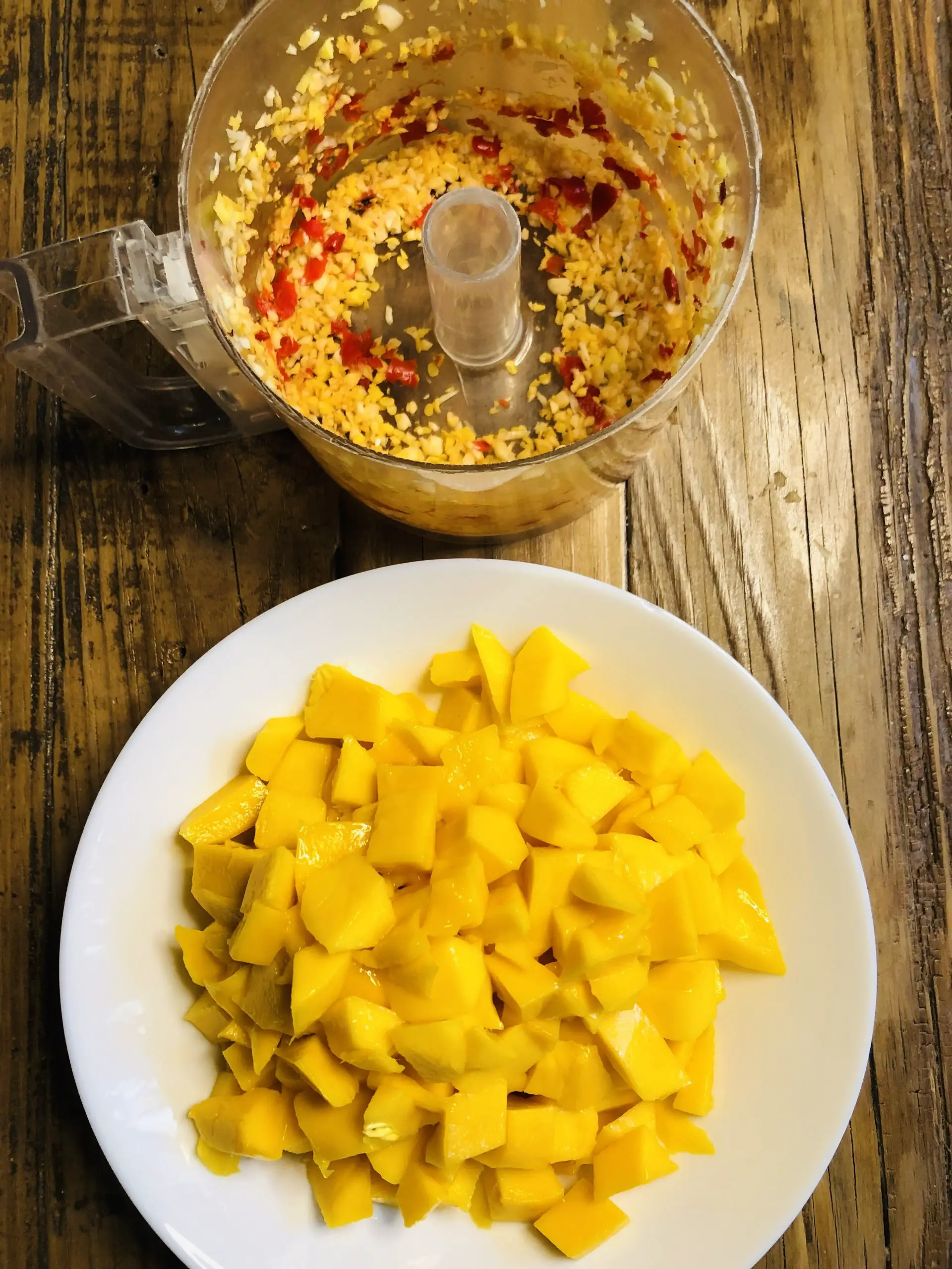 Pieces of cut up mango in a white bowl, minced chilies, and garlic in a food processor.