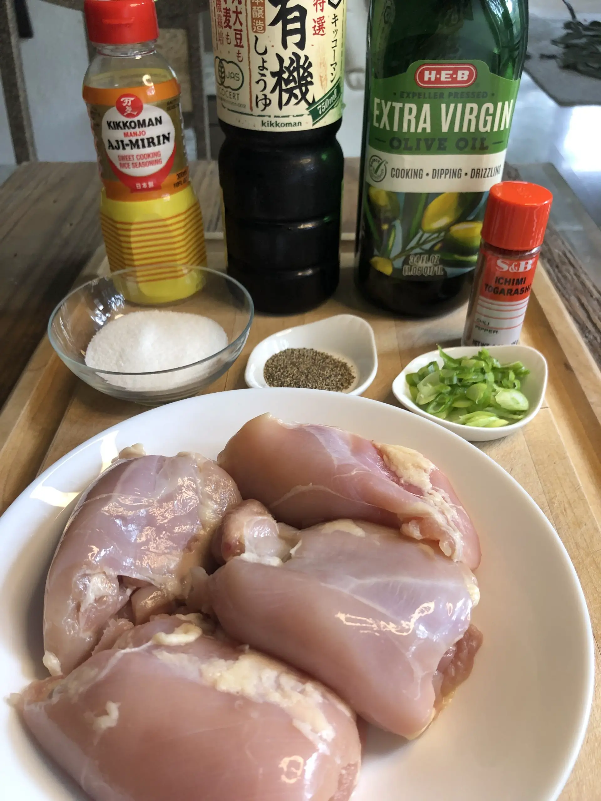 Chicken thighs in a white bowl, Aji-Mirin bottle, bottle of soy sauce, olive oil bottle, sugar in a glass container, pepper in a small dish, green onions in a white dish, Ichimi Togarashi.