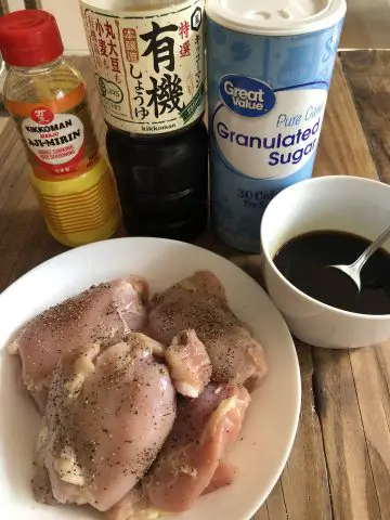 Chicken thighs with black pepper in a white bowl, teriyaki sauce in a white bowl, Aji-Mirin bottle, bottle of soy sauce, and a container of sugar.