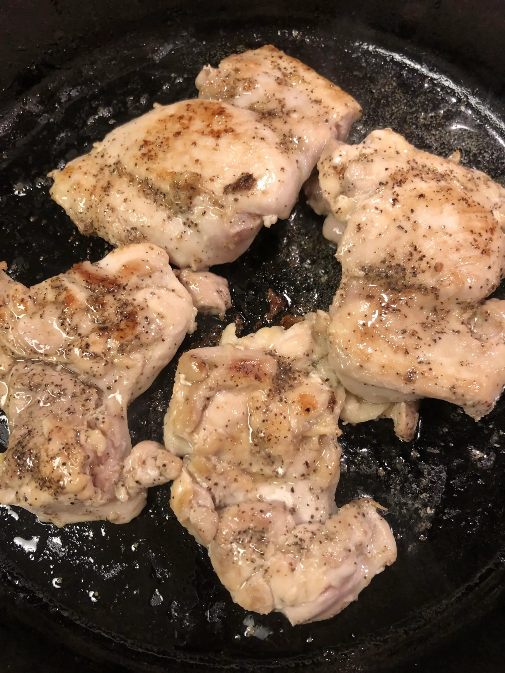 Chicken thighs frying in a cast iron skillet seasoned with pepper and starting to brown.